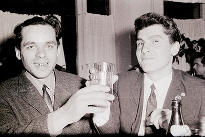 Two men in formal attire pose indoors, likely mid-20th century (1950s–1960s). One holds a wineglass, the other a bottle with ...