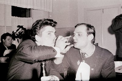 Two men in formal attire share a playful toast at an indoor event, likely mid-20th century. One pours from a bottle into the ...
