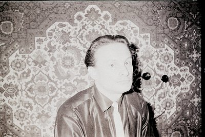 Portrait of a man in mid-20th century formal attire—dark suit, white shirt, and tie—against an ornate patterned wallpaper bac...