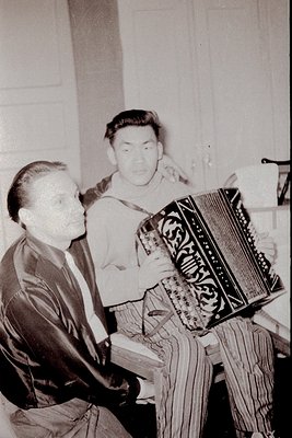 Two men in 1960s indoor setting playing ornate accordion. Formal attire suggests cultural event or performance. Ornate instru...