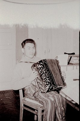 Vintage black-and-white photo of a man seated playing a decorated accordion, likely mid-20th century. Indoor setting with woo...
