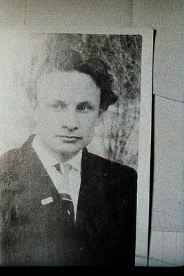 Black-and-white portrait of a man in formal attire: dark suit, white shirt, and tie with a pin. The hairstyle and clothing su...