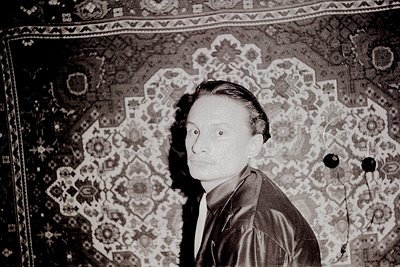 Portrait of a man in mid-20th century attire, seated against an ornate patterned rug with geometric floral motifs. Dark suit ...