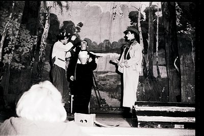 Vintage black-and-white stage performance with three actors in period costumes: a woman in a high-collared dress, a man in a ...