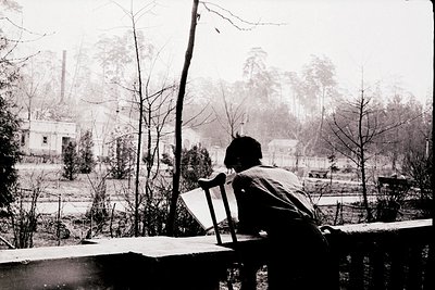 A lone figure sits on a wooden bench overlooking a misty, industrial landscape. The person faces away, wearing a long coat an...