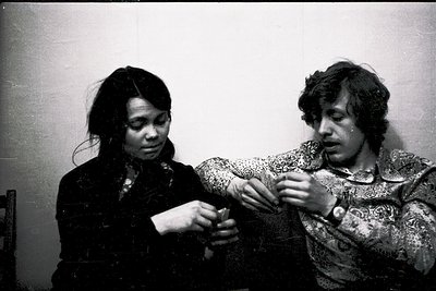 Black-and-white candid of two individuals adjusting a wristwatch in a dimly lit interior, likely 1970s–1980s. The woman wears...