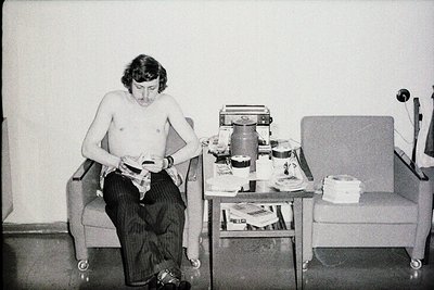 Shirtless man seated in a wheeled armchair, holding a small handheld device (likely a tape recorder or early audio player) in...