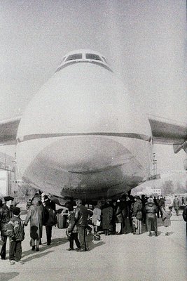 Vintage black-and-white photo of a **Tupolev Tu-114** (Soviet-era airliner) with passengers boarding via external stairs. Cro...