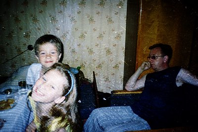 Vintage indoor portrait featuring a man and two children in a dimly lit room with floral wallpaper. The man wears glasses and...
