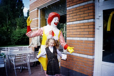 A 1970s-era McDonald’s mascot (Ronald McDonald) poses with a young girl in a matching costume outside a brick-façade restaura...