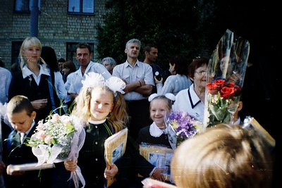 School graduation ceremony in Eastern Europe, likely 1980s–1990s. Girls in traditional white headbands and black dresses hold...