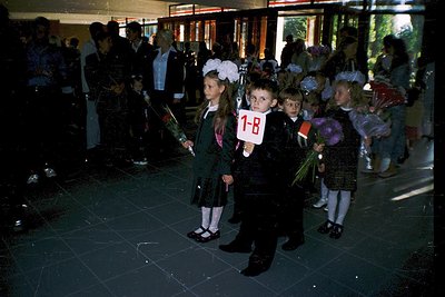 First-day-of-school ceremony in a Soviet-era school hallway, likely Eastern Bloc (1970s–1980s). Children in formal uniforms h...