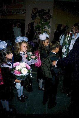 Schoolchildren in formal attire—girls in white blouses, dark skirts, and floral headbands holding bouquets, boys in suits—gat...
