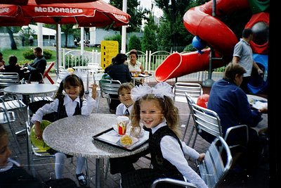 Vintage McDonald’s playground scene with children in 1970s-80s school uniforms (blazers, ties, headbands) seated at metal tab...