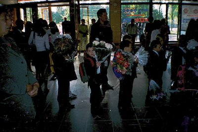 Crowded indoor hallway with students in formal attire holding floral bouquets, likely a graduation ceremony. Concrete pillars...