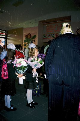 A group of schoolgirls in 1970s Eastern European attire—white headscarves, floral bouquets, and dark skirts—stand in a dimly ...