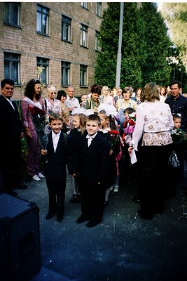 A group of children in formal attire—jackets, ties, and white bows—stands in front of a Soviet-era brick building with narrow...