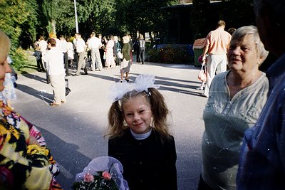 Vintage outdoor gathering with a young girl in a black dress and white headband, flanked by an elderly woman in a light-color...