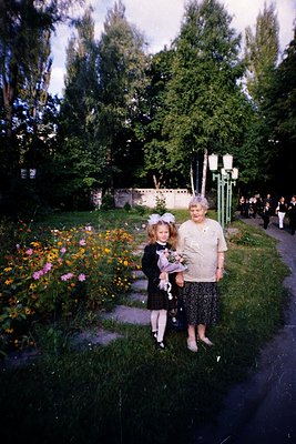 Vintage garden portrait featuring a woman in 1960s-era dress and a young girl in formal attire, holding a bouquet. Lush green...