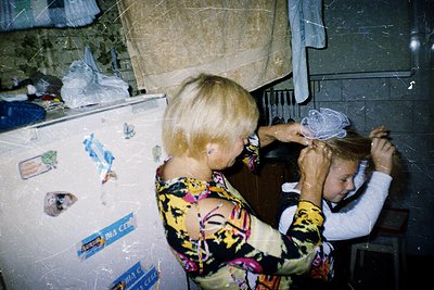 A woman in a floral-patterned blouse assists a child with a hairnet in a modest kitchen. Fridge covered in magnets and sticke...