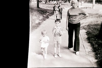 Black-and-white street scene from the 1960s–70s: woman in high-waisted trousers and sweater escorts two children (one in stri...