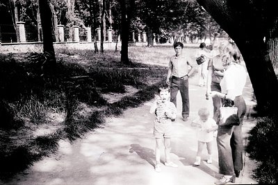 Vintage black-and-white photo of a family strolling in a park, likely mid-20th century. Adults in button-down shirts and knee...