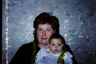Vintage sepia-toned photo of a woman holding a baby, likely from the 1970s–1980s. The baby wears a blue garment with gold emb...