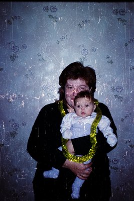 A woman holds a baby adorned with a yellow floral lei against a floral-patterned wall, likely from a 1970s-1980s indoor setti...