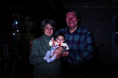 A family portrait featuring an elderly couple holding a baby indoors, likely during the holiday season. The woman wears a tex...