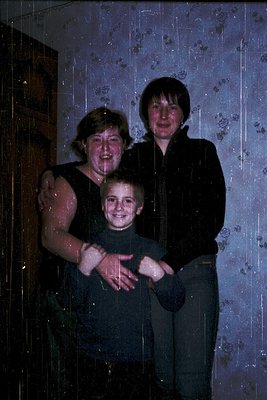 Family portrait taken indoors, likely in a residential setting during the 1970s–1980s. Three individuals pose in a dimly lit ...