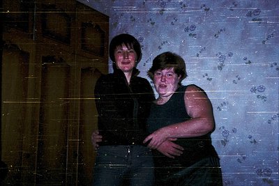 Two individuals pose indoors against a wallpapered backdrop featuring floral patterns. The woman on the left wears a dark lon...