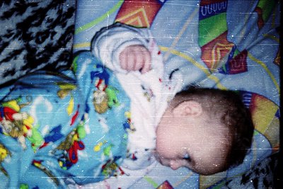 Vintage-style photo of an infant wrapped in a colorful, cartoon-themed blanket with visible wear. The child appears relaxed, ...