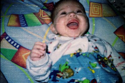 Vintage-style photo of an infant laughing while holding a yellow toy. Patterned blue blanket with geometric and animal prints...