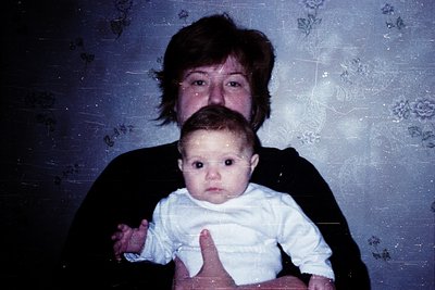 Vintage portrait of an adult holding a baby indoors, likely mid-1980s–1990s. Floral wallpaper and soft lighting suggest a dom...