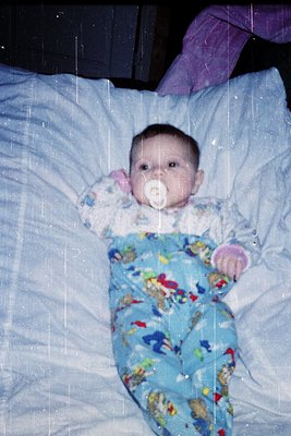 Candid shot of an infant in a blue floral-patterned sleeper, lying on a light blue blanket with a pacifier. Soft lighting sug...