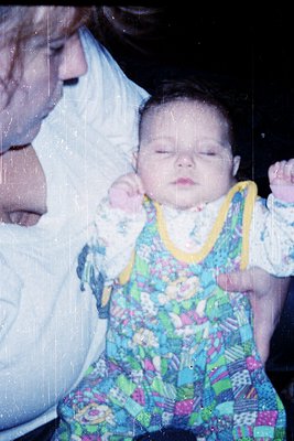 Tender moment of an infant in a floral-patterned romper with yellow straps, cradled by an adult in a light-colored blouse. So...