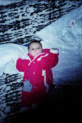 Vintage photo of a toddler in a red snowsuit with a blue elephant patch, posing indoors near a snow-covered igloo-like struct...