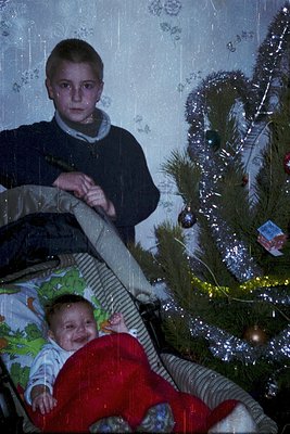 Vintage holiday photo: boy (1970s-style sweater vest) leans over a baby in red sleeper, seated on a couch beside a decorated ...
