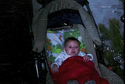 Vintage holiday photo of an infant in a sleigh-style carrier, wrapped in a floral-patterned blanket and red cloth. Snowflakes...