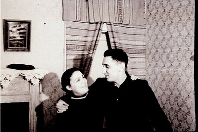 Vintage black-and-white portrait of a man and woman in an indoor setting, likely mid-20th century. The man wears a dark unifo...