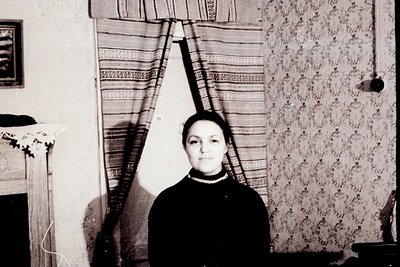 Vintage black-and-white portrait of a woman standing indoors, framed by striped curtains and floral wallpaper. She wears a da...