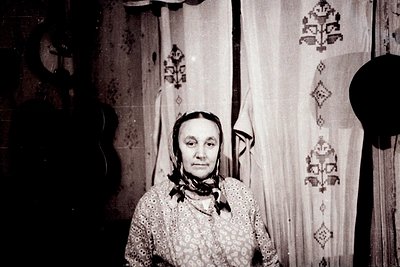 Vintage black-and-white portrait of a woman in traditional embroidered attire, seated indoors with patterned fabric curtains ...