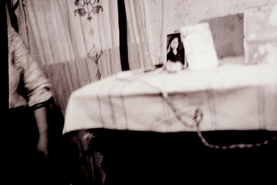 Vintage black-and-white interior shot of a bedroom with a single bed draped in a white sheet. A framed photo on the bedside t...