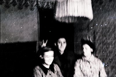 Vintage black-and-white portrait of three individuals indoors, likely mid-20th century. The central figure poses with a peace...
