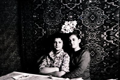 Two young women pose indoors against an ornate patterned wall, likely mid-20th century. The woman on the right wears a floral...