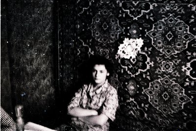 Vintage black-and-white portrait of a young woman seated against an ornate patterned wallpaper featuring geometric floral mot...