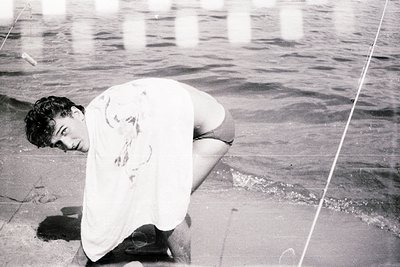 Black-and-white shot of a man kneeling on sandy beach, arms resting on knees, facing water. Distinctive 1960s–70s beachwear: ...