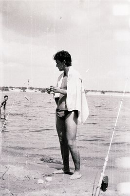 Vintage black-and-white beach scene featuring a man in minimalist swimwear (shorts and draped towel) holding a fishing rod, s...