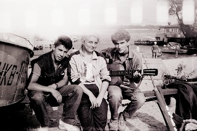 Three young people pose outdoors in a 1970s-era setting, likely Eastern Europe. The man on left wears a dark vest and jeans; ...