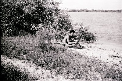 Black-and-white lakeside scene featuring two individuals seated on sandy shore, surrounded by reeds and greenery. Mid-20th ce...